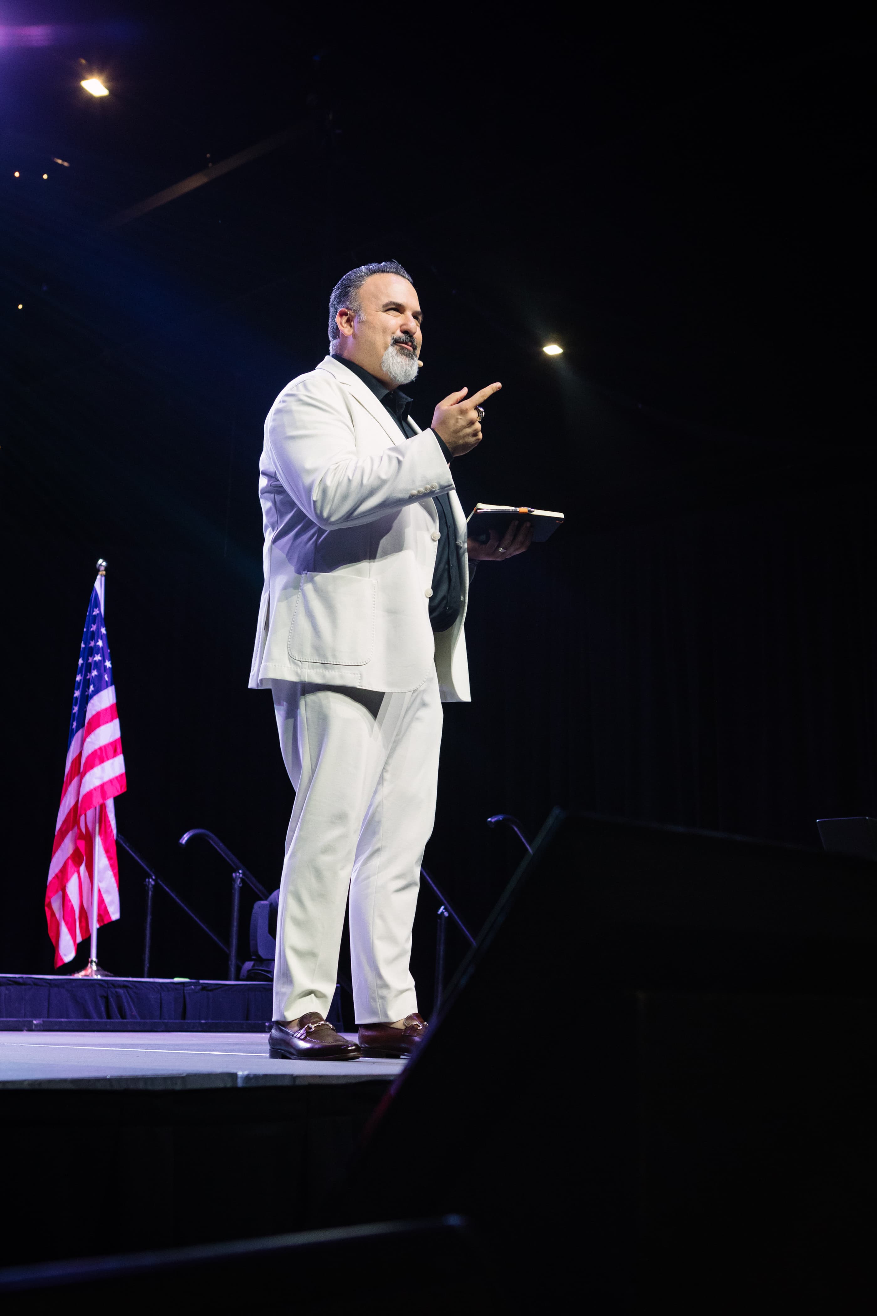 Frank Delio in a white suit speaking on stage